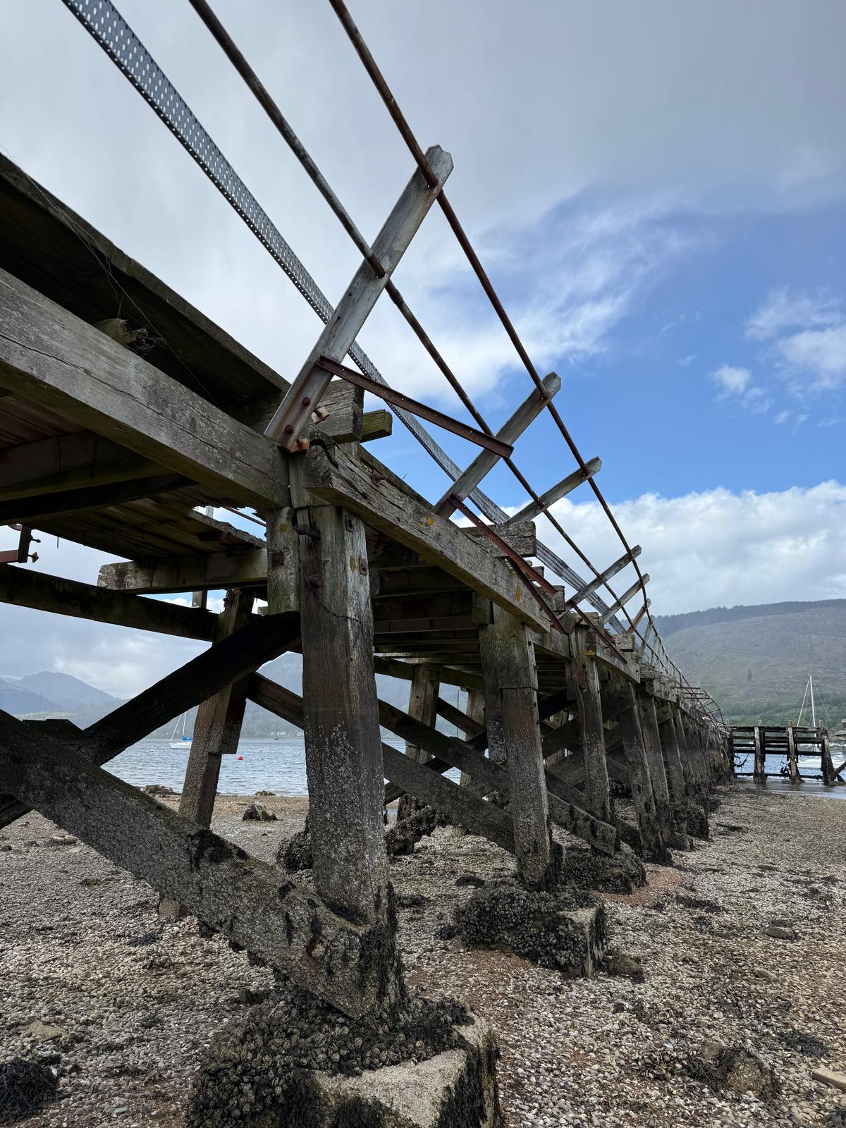 Side view of Ardnadam Pier's weathered timber substructure with steel bracing, photographed at low tide on the Holy Loch.
