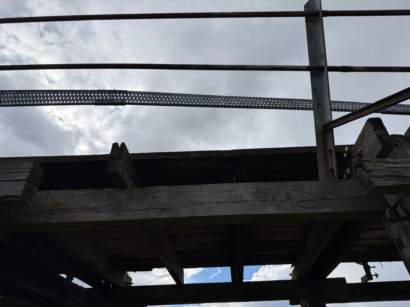 Underside of the pier deck showing cracked timber beams and perforated steel bracing against an overcast sky.