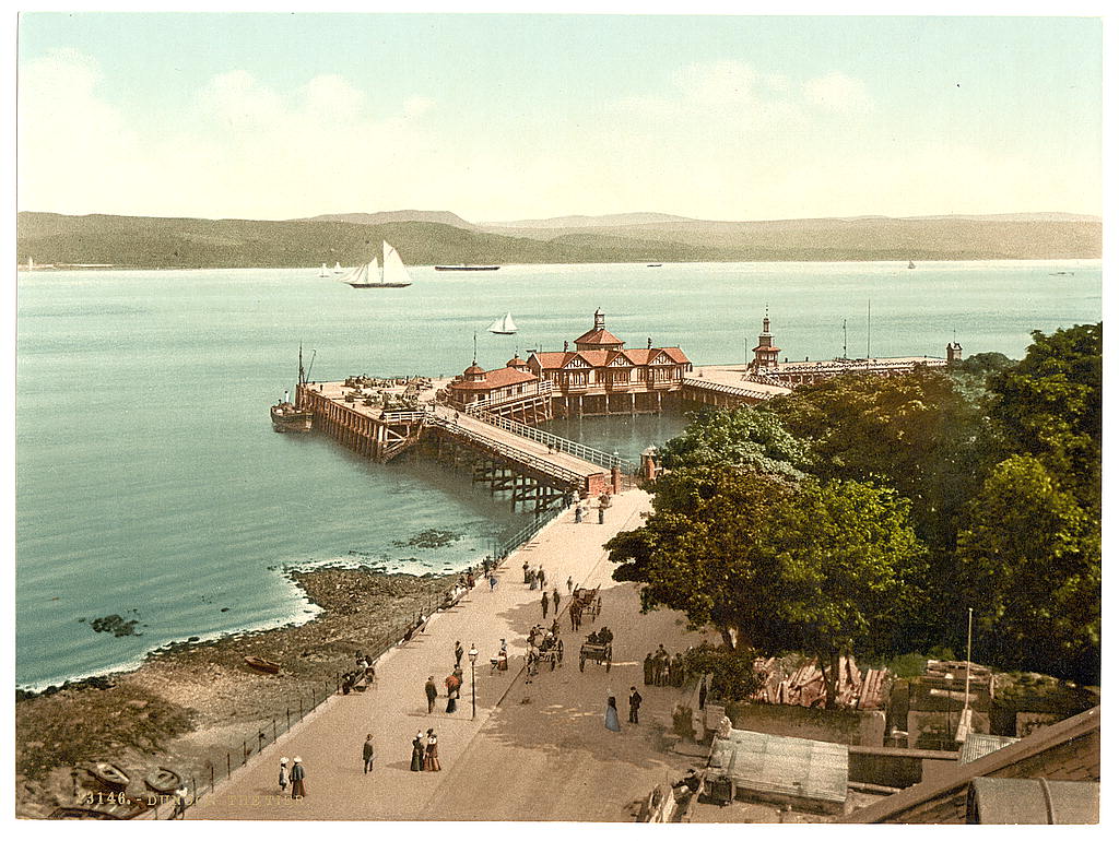 Photochrom of the wooden pier at Dunoon with promenading visitors and moored paddle steamers.