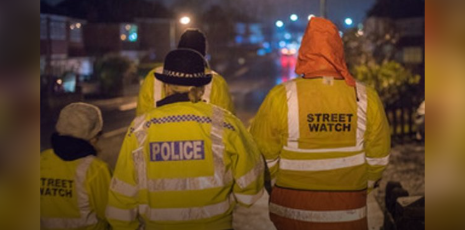 Two Streetwatch volunteers in high-visibility jackets walking a Glasgow street at night alongside a police officer.